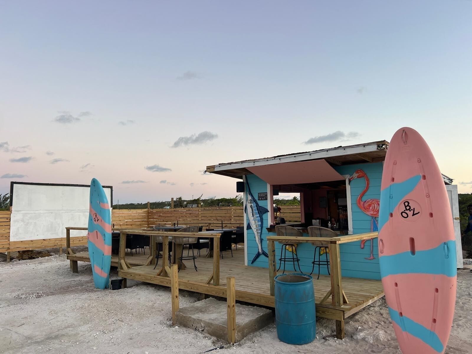 A beachside bar with a wooden deck, outdoor seating, and surfboards.
