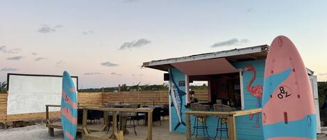 A beachside bar with a wooden deck, outdoor seating, and surfboards.