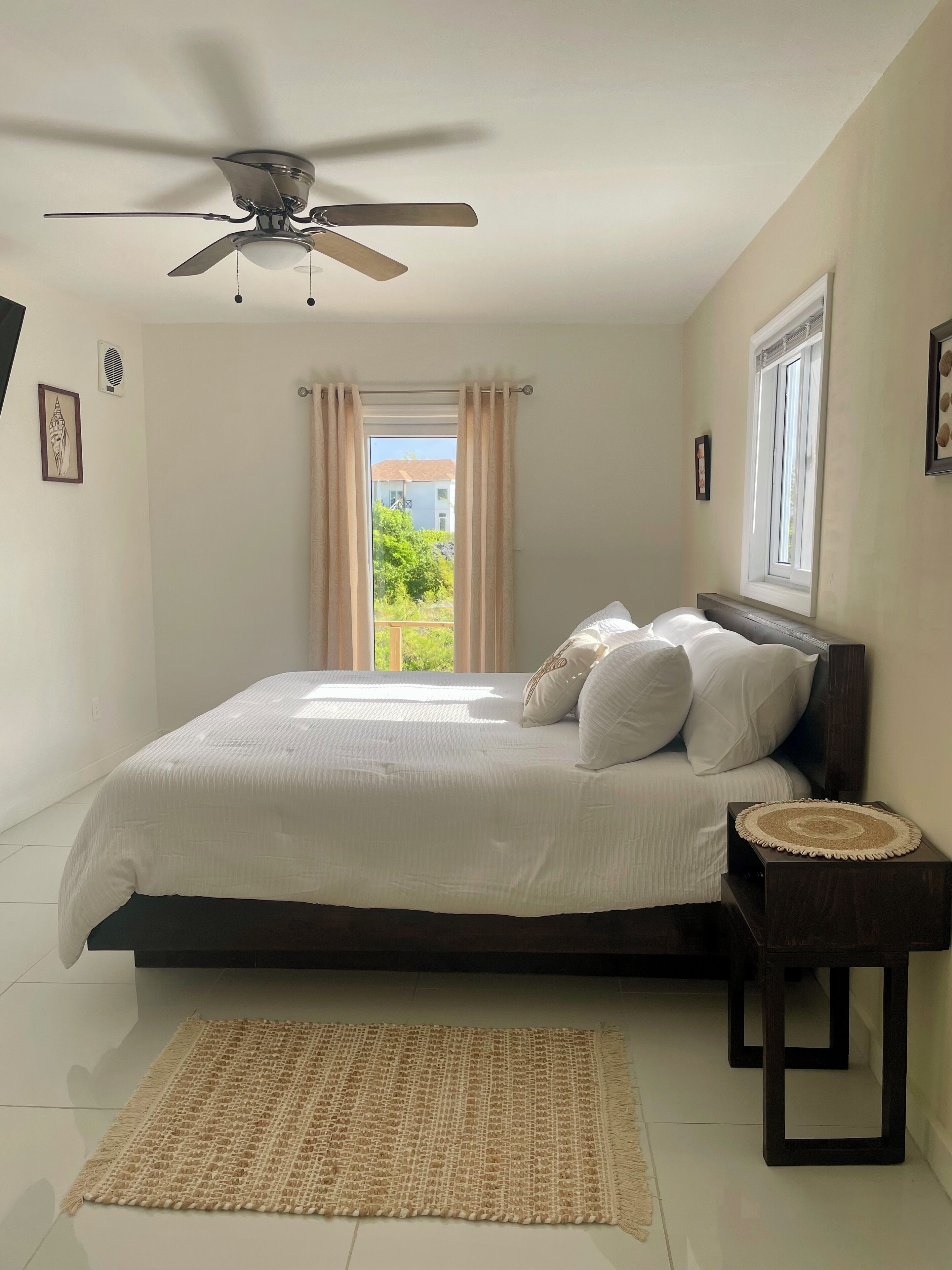 Une chambre à coucher avec un lit, un ventilateur de plafond, une fenêtre avec des rideaux, une table de chevet et une vue sur la verdure à l’extérieur.