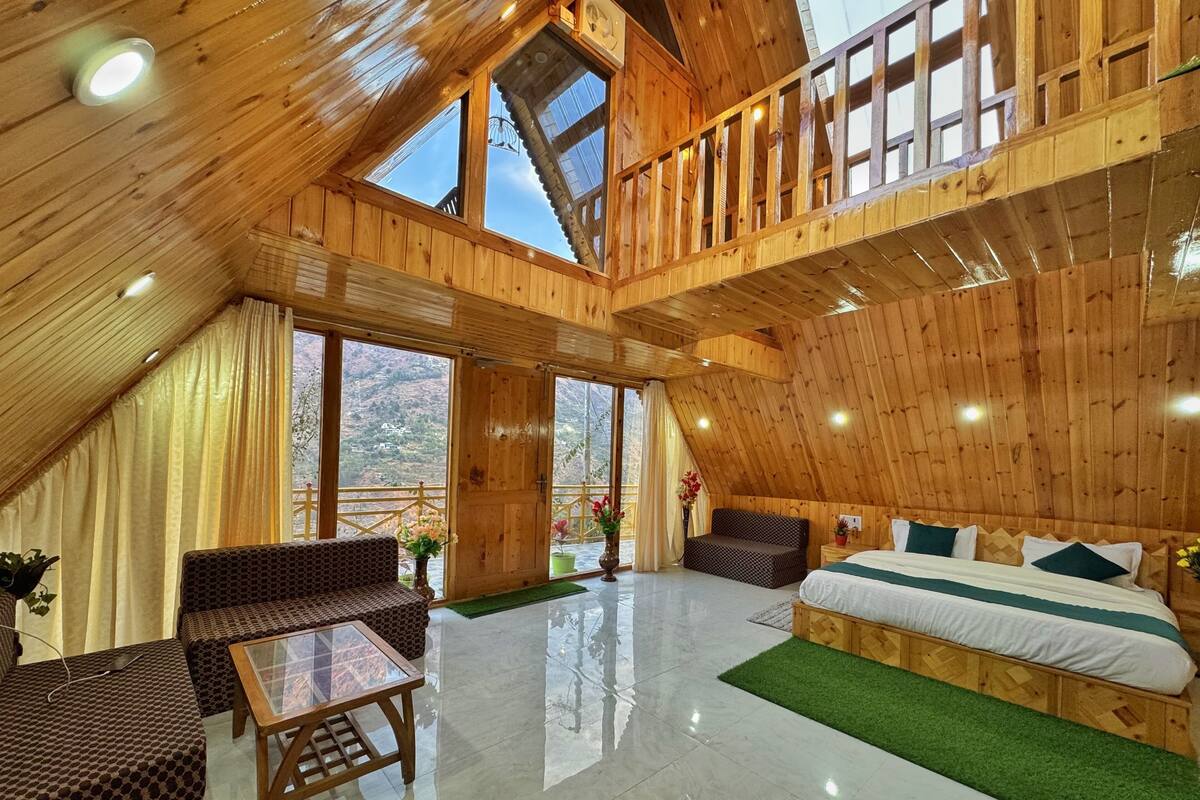A spacious room with a high ceiling, wooden walls, a balcony, and a bed with a green bedspread.