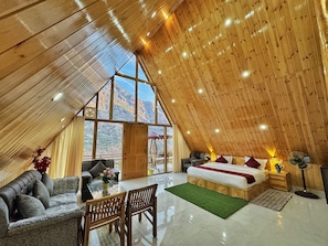A spacious room with a high ceiling, a large bed, a seating area with a table, and a view of mountains through large windows.