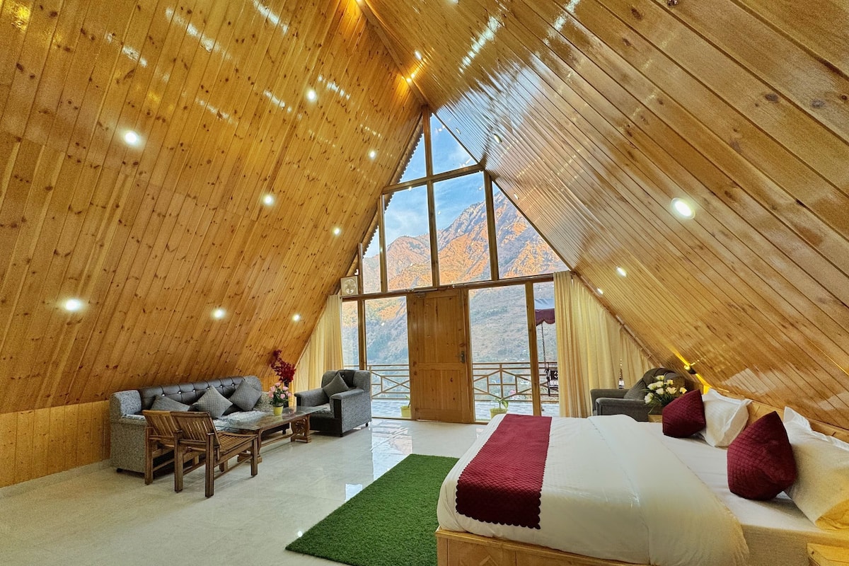 A spacious room with a high ceiling, wooden walls, a large bed, a sofa, and a view of mountains through large windows.