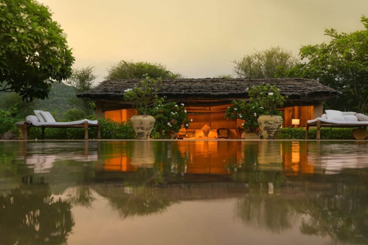 Un bâtiment traditionnel doté d’un toit de chaume, entouré d’une végétation luxuriante et bordé d’une étendue d’eau calme dans laquelle se reflète la structure.