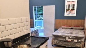 A compact kitchen with a coffee maker, sink, and microwave. There is a bed with a mattress and pillows, and a window with a view of greenery. - Marmeu Enseada (Guarujá)