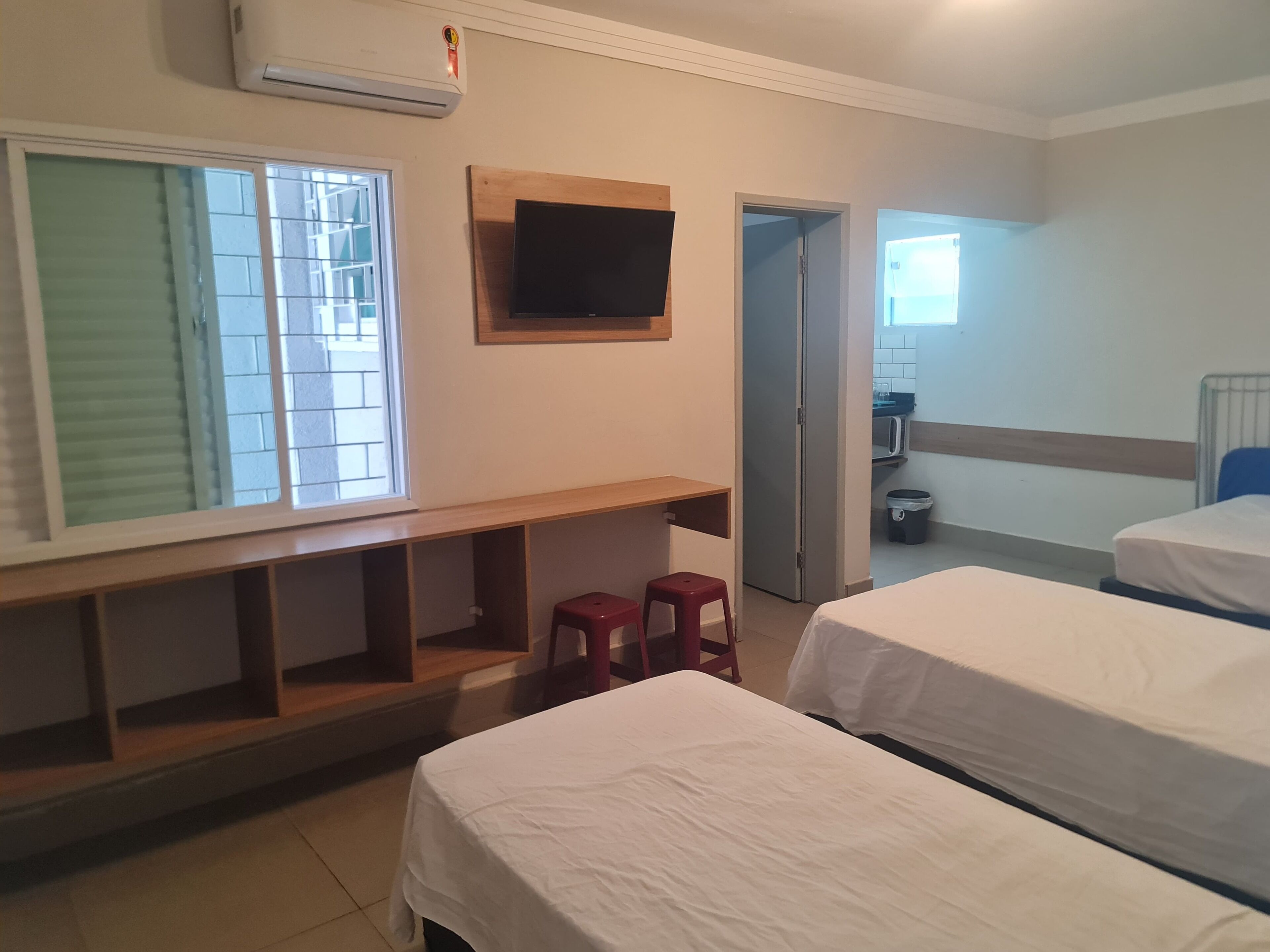 Une chambre d’hôtel avec deux lits, un téléviseur fixé au mur, une fenêtre avec des stores et, au fond, une petite cuisine.