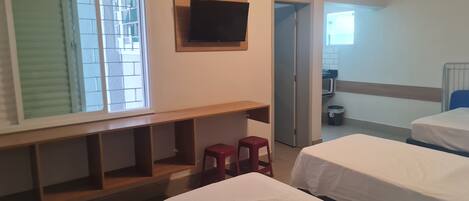 Une chambre d’hôtel avec deux lits, un téléviseur fixé au mur, une fenêtre avec des stores et, au fond, une petite cuisine.