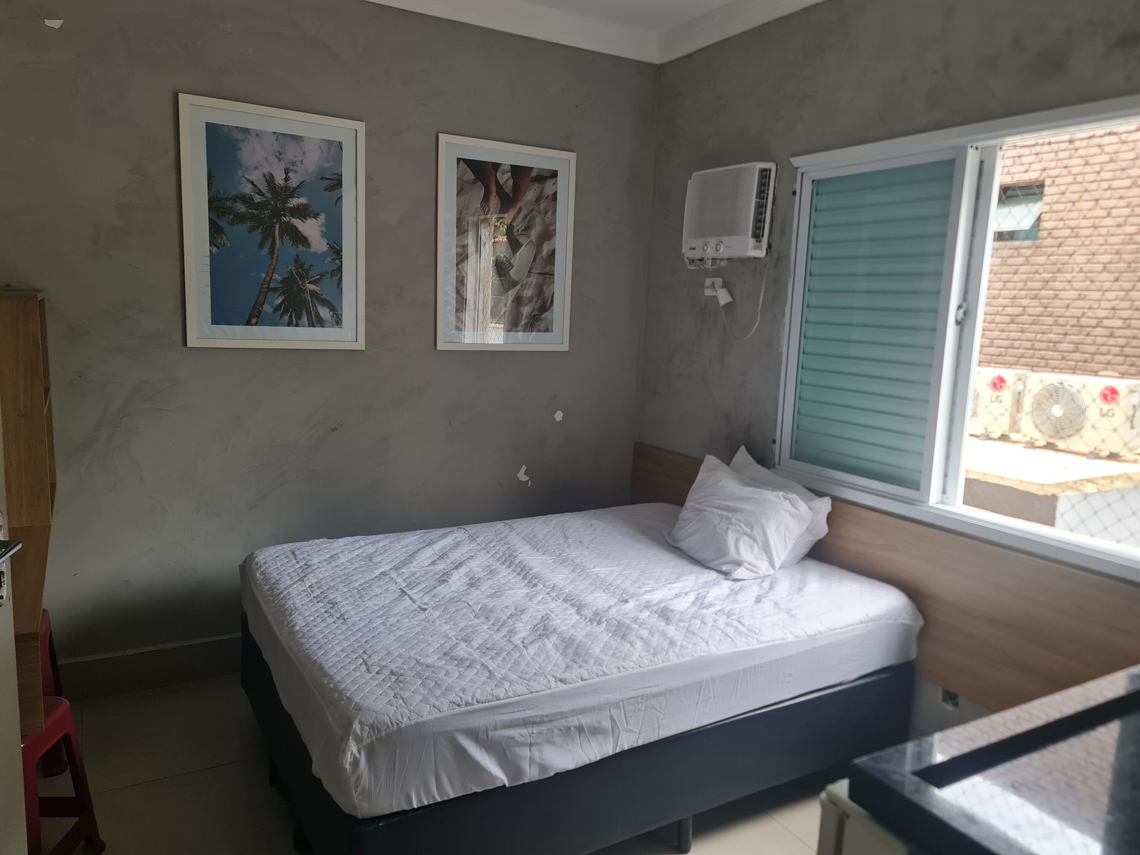 Un dormitorio con cama, dos cuadros en la pared, aire acondicionado y una ventana con persianas.