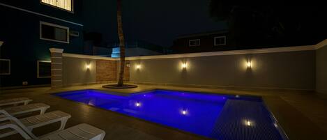 Piscina exterior con sillones de descanso y una palmera, rodeada de muros con iluminación exterior.