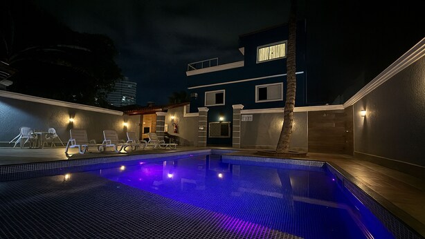 A nighttime swimming pool area with lounge chairs and a building in the background. - Marmeu Enseada (Guarujá)