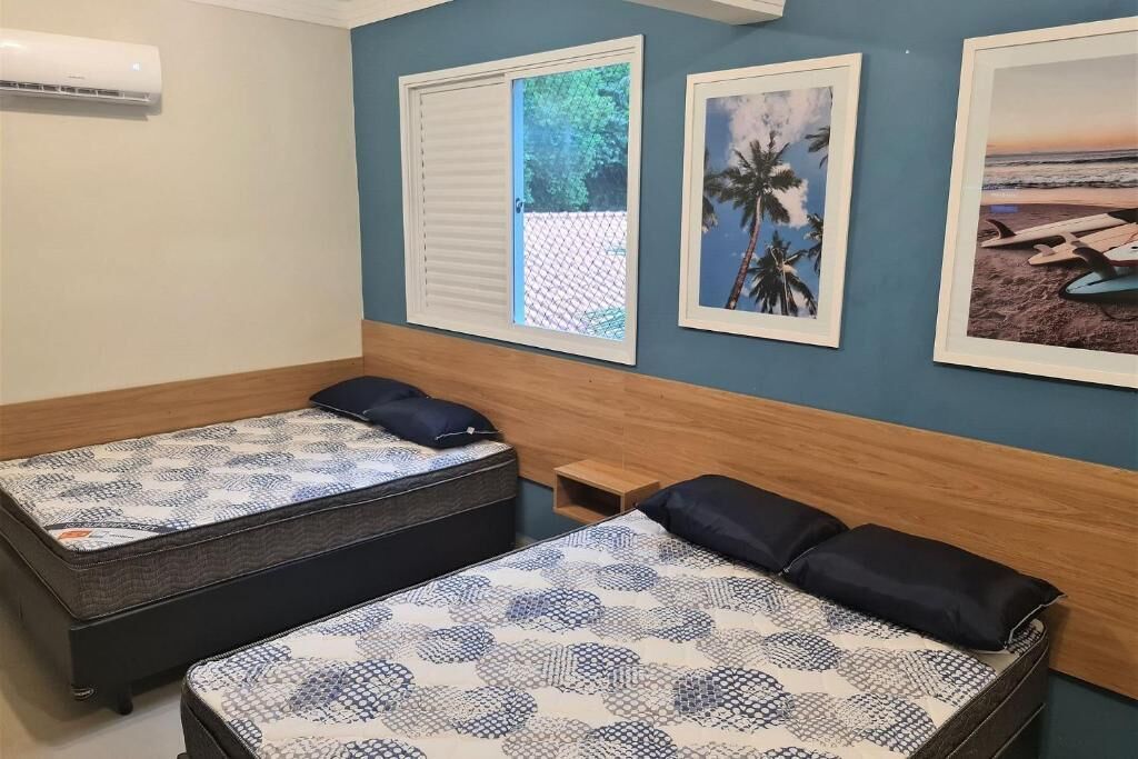 Quarto com duas camas, janela com vista para vegetação e duas imagens emolduradas na parede.