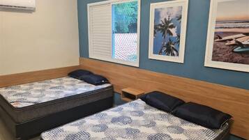 Quarto com duas camas, janela com vista para vegetação e duas imagens emolduradas na parede.
