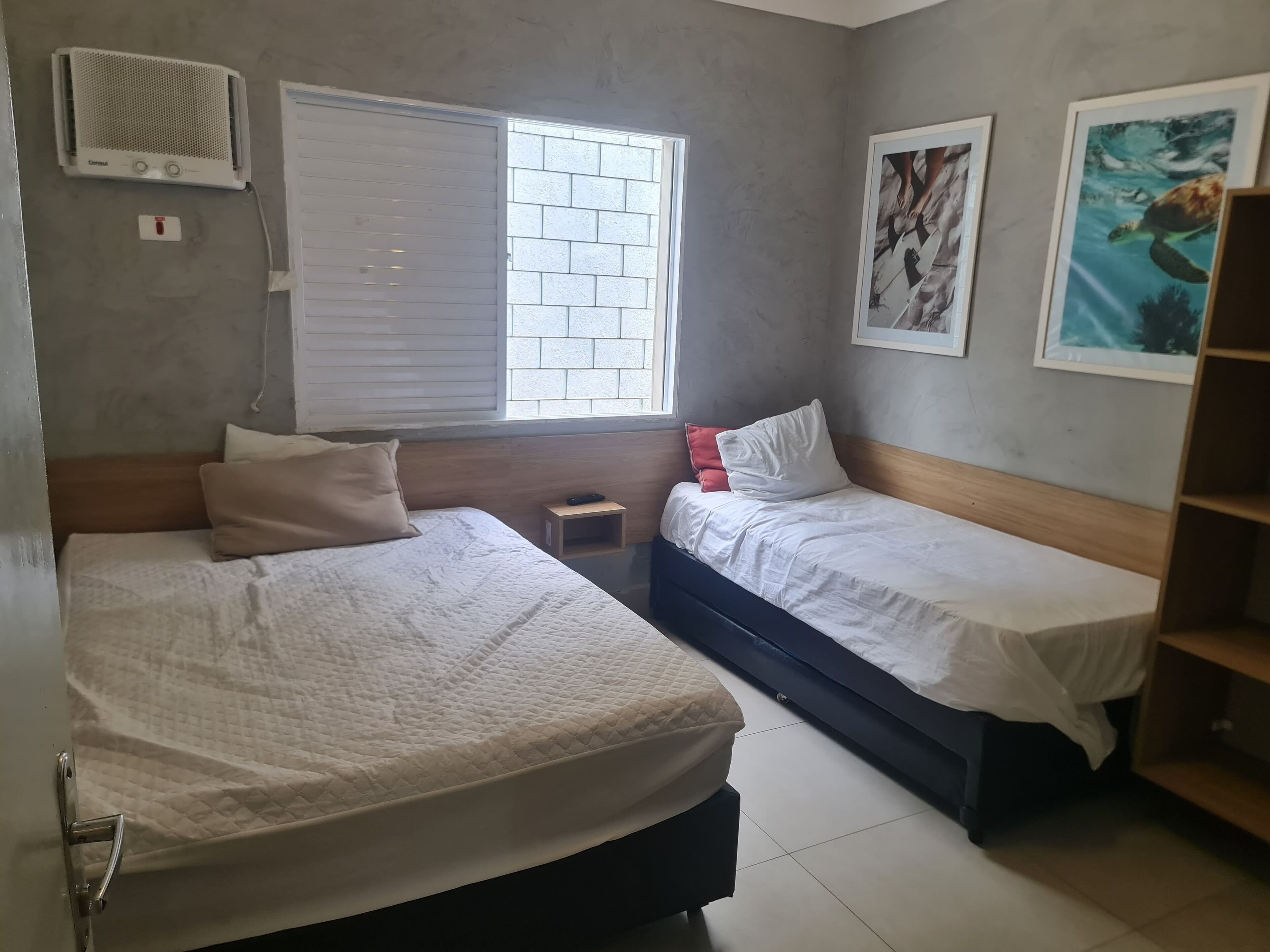 A room with two beds, a window with blinds, a small bedside table, and a wall-mounted air conditioner.