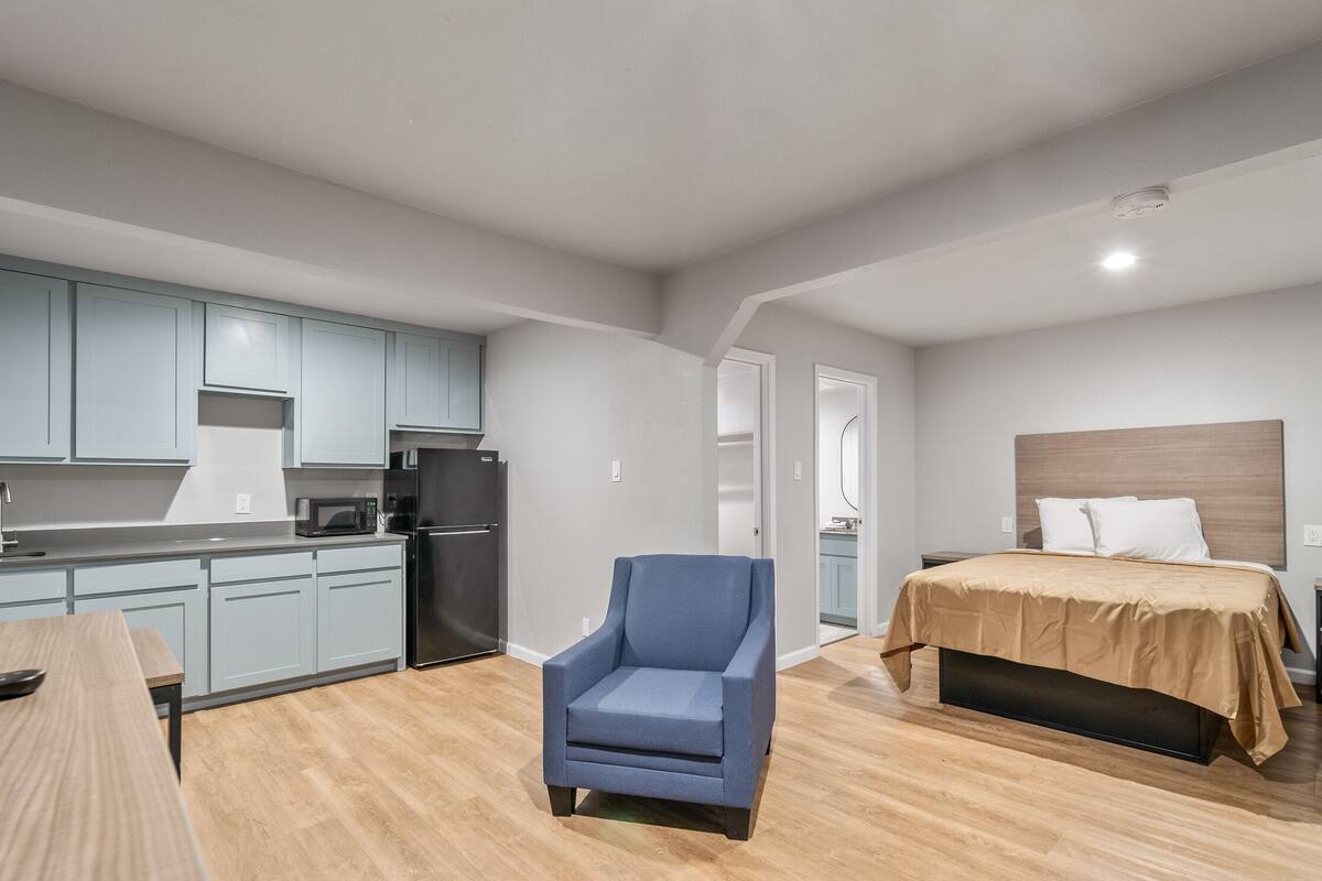 A compact hotel room with a kitchenette, a single bed, a blue armchair, and a wooden table.