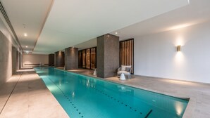 A modern indoor swimming pool with clear lanes, large pillars, and a seating area with a chair and table. - Melbourne Corporate Stays – Docklands Waterfront (Docklands)