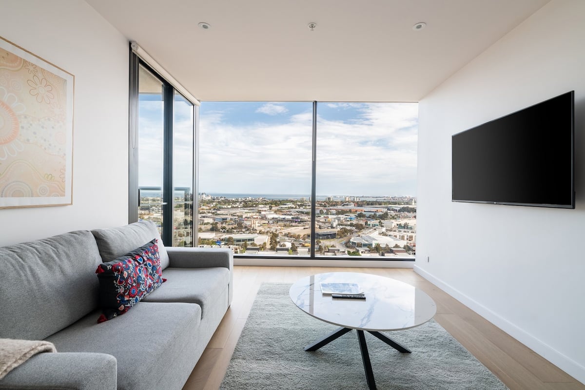 Een moderne woonkamer met een groot raam, een grijze bank, een ronde salontafel en een flatscreen televisie.