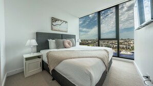 A modern bedroom with a large bed, a nightstand, a lamp, and a city view through large windows. - Melbourne Corporate Stays – Docklands Waterfront (Docklands)