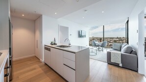 A modern kitchen with a central island, a dining area with a sofa and chairs, and a large window offering a city view. - Melbourne Corporate Stays – Docklands Waterfront (Docklands)