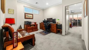 A spacious living room with a leather sofa, wooden coffee table, and a desk with a computer. A large window provides natural light, and a door leads to an outdoor area. - StayAU Pakenham Townhouse with Lakeview (Pakenham)