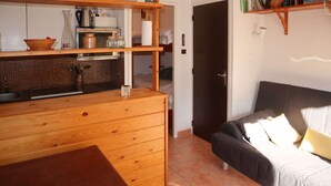 Une cuisine de taille réduite, dotée de meubles en bois, d’un plan de travail et d’un évier. Un canapé avec des coussins est placé près d’une porte qui donne sur une autre pièce.