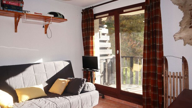 Un salon avec un canapé gris, un téléviseur posé sur un meuble, une étagère en bois et une porte-fenêtre coulissante donnant sur un balcon.