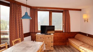 A cozy room with a wooden table, chairs, a sofa, a TV on a wooden cabinet, and large windows with curtains. - APARTMENT N° 706 LE BELVEDERE (LES ORRES 1650)