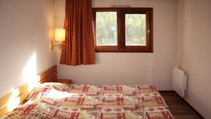 A bedroom with a wooden bed, a window with curtains, a wall-mounted lamp, and a radiator. - APARTMENT N° 706 LE BELVEDERE (LES ORRES 1650)