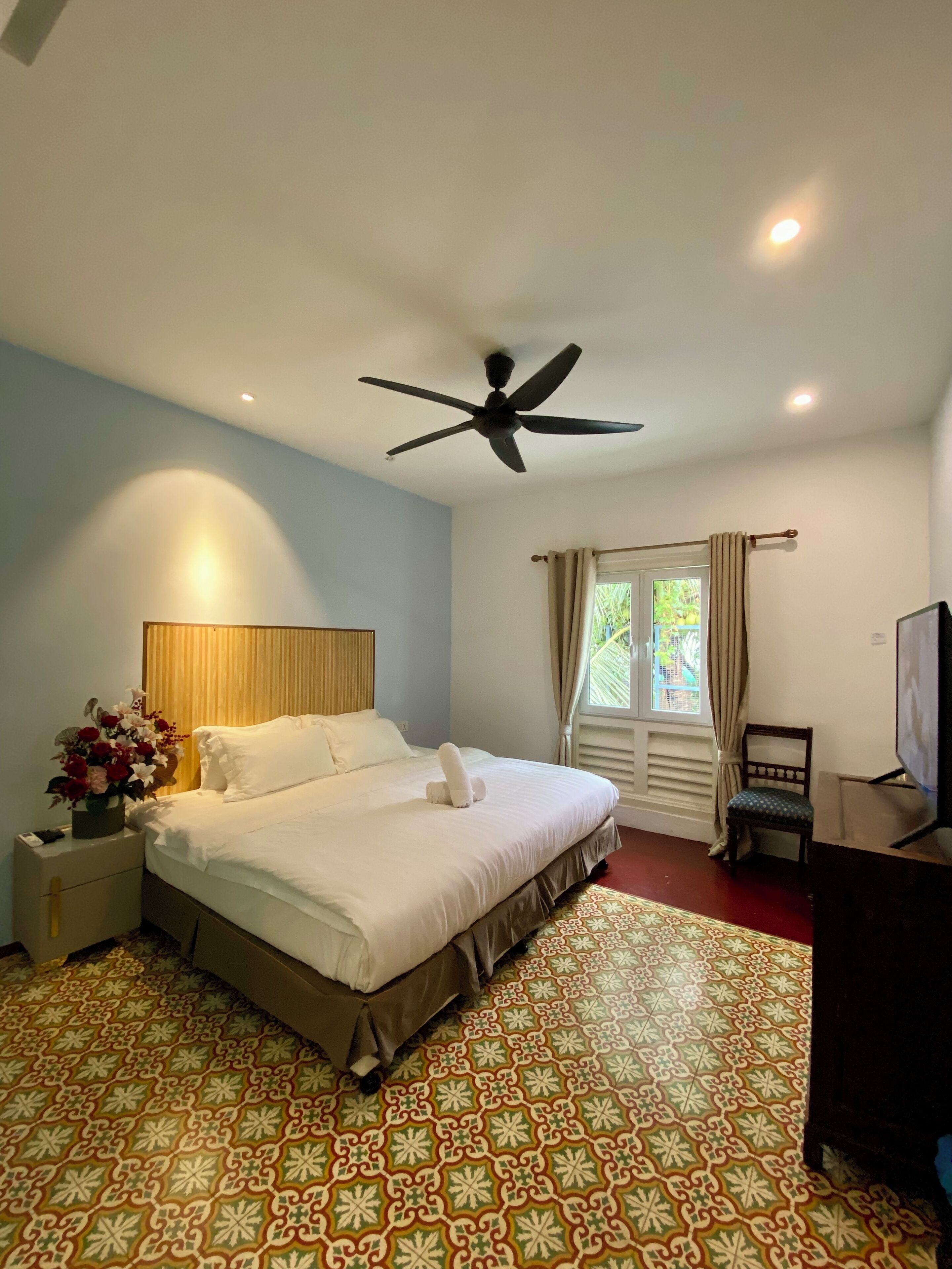 A hotel room with a bed, a ceiling fan, a nightstand with a vase of flowers, a window with curtains, and a television.