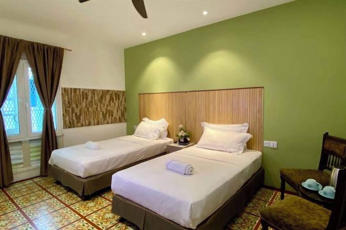 A hotel room with two single beds, a small table with a chair, and a window with curtains.