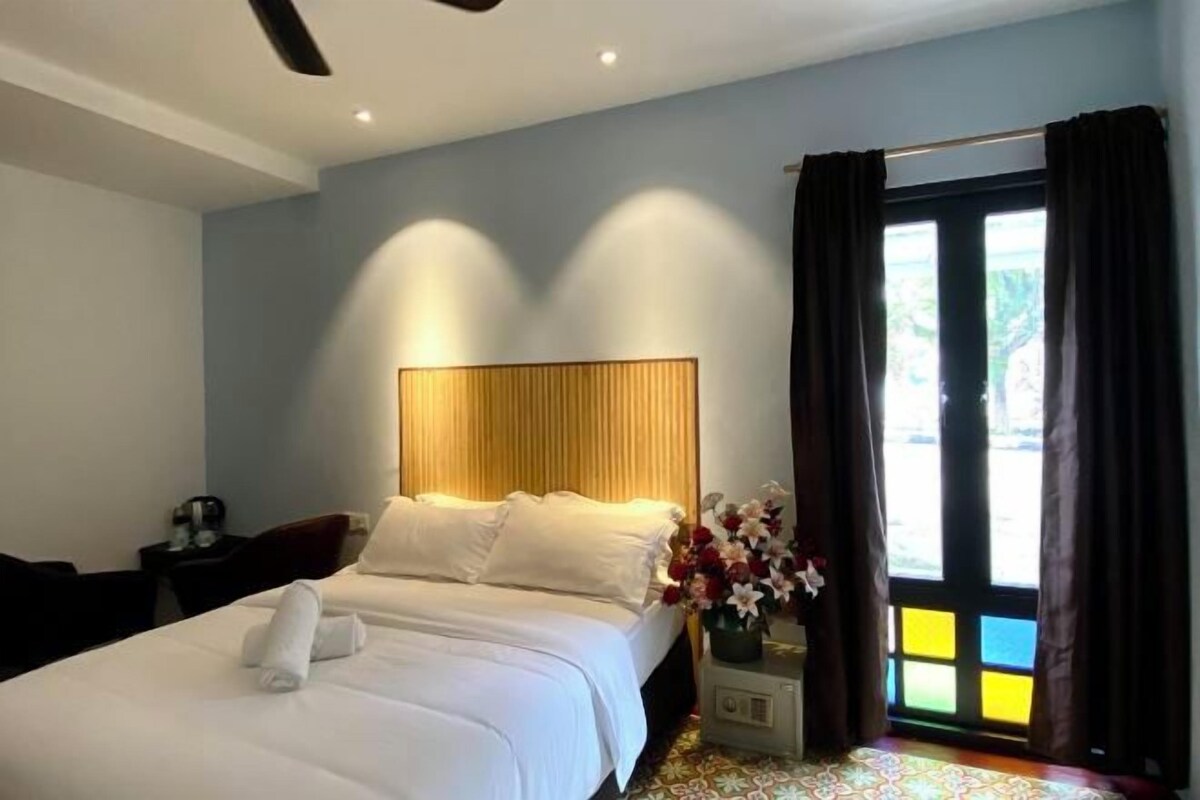 A neatly made bed with white linens and a rolled towel, a bedside table with a lamp and a vase of flowers, and a window with curtains.