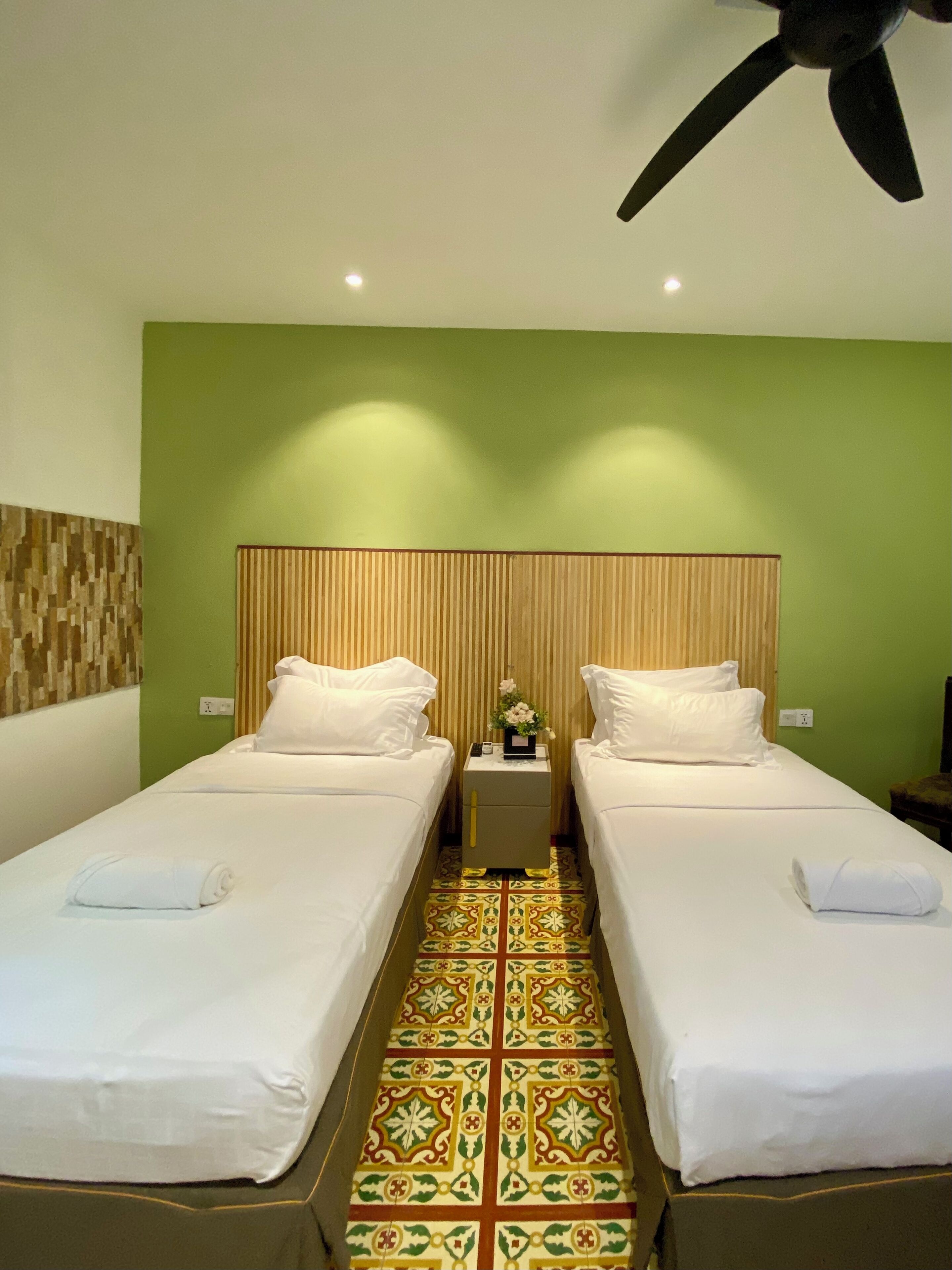 A hotel room with two single beds, a green accent wall, a patterned floor, and a ceiling fan.