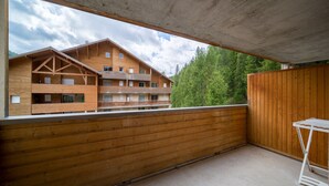 Un balcón de madera con techo de hormigón, que da a un edificio de varios pisos con balcones y vistas a árboles verdes.