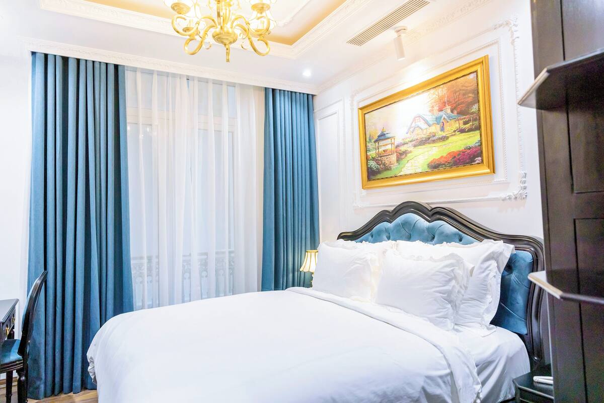 Une chambre d’hôtel avec un grand lit, un lustre, un tableau au mur et des rideaux bleus.