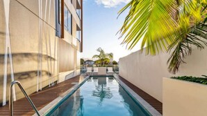 Une piscine dotée d’une terrasse en bois, entourée de bâtiments modernes et de palmiers.
