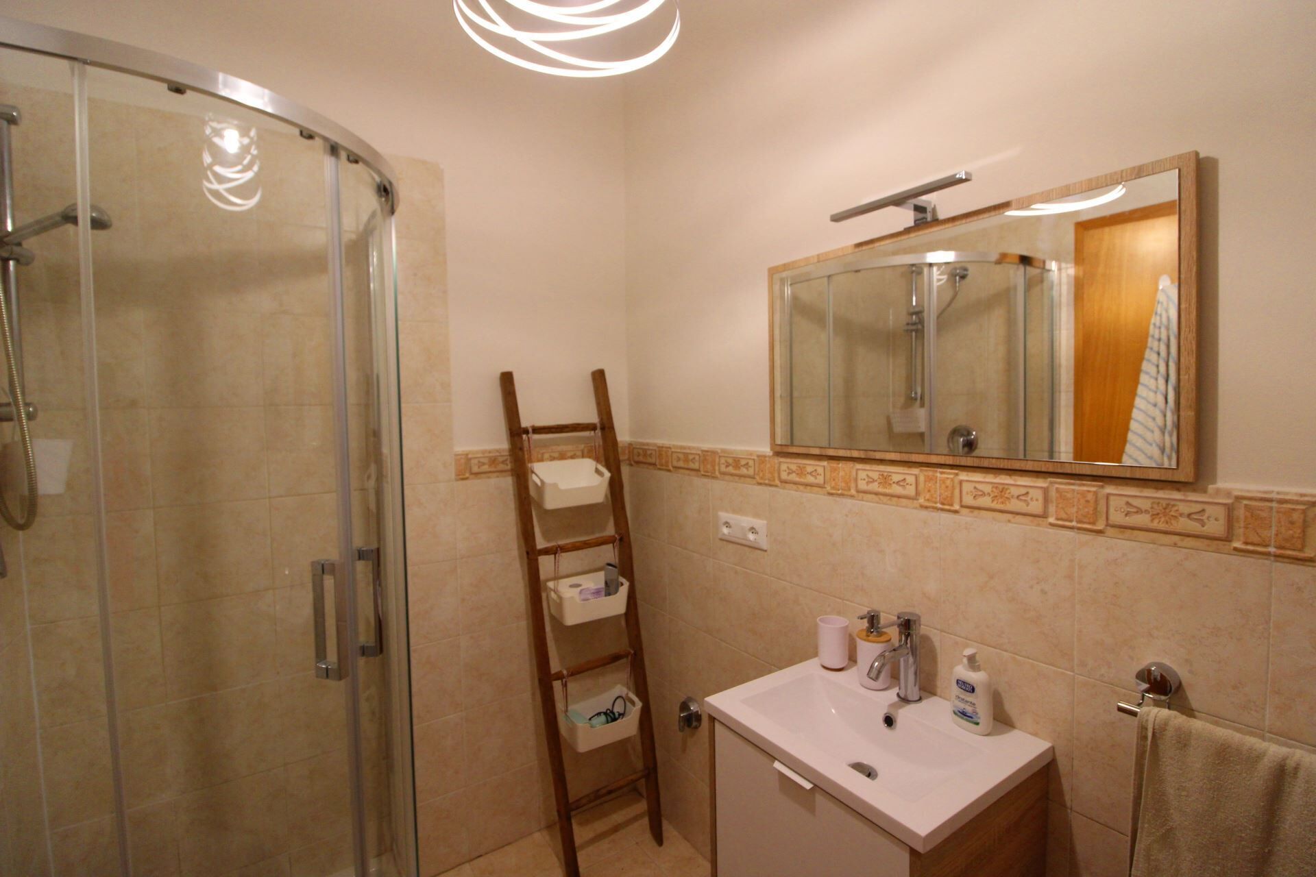 A bathroom with a glass shower enclosure, a wooden ladder, a sink with toiletries, and a mirror above the sink.