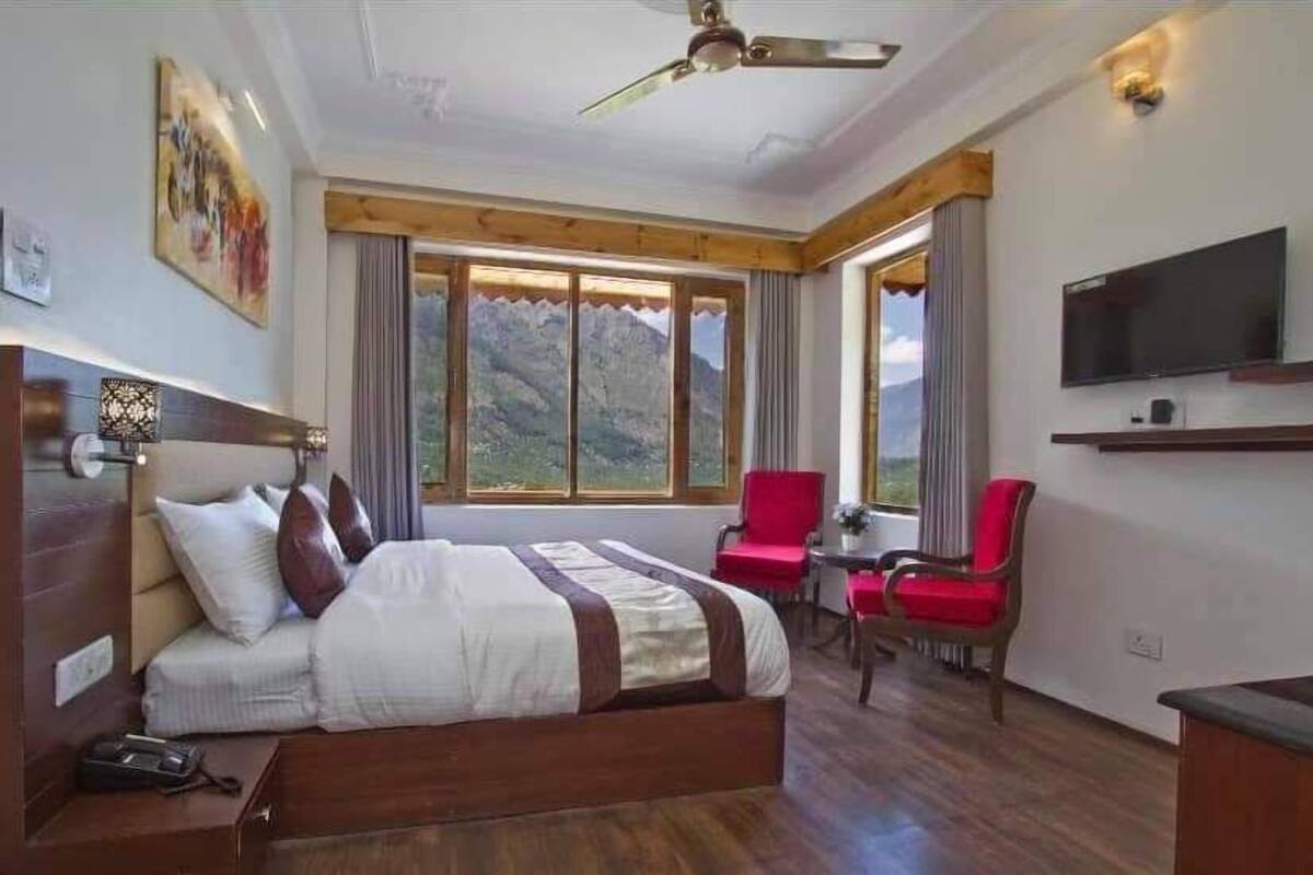Habitación de hotel con una cama grande, un escritorio, una silla, un televisor y vistas a las montañas a través de las ventanas.