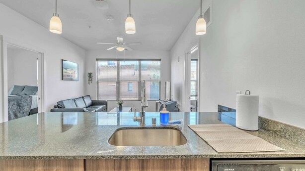 A modern kitchen with a granite countertop, stainless steel sink, and an AMANA dishwasher. There is a ceiling fan, pendant lighting, and a view of the outside through large windows. - Big Flat + 2 min to UM + 95 Walkscore (Missoula)