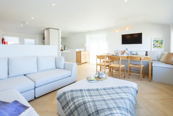 A modern living room with a sofa, a dining table with chairs, a flat-screen TV mounted on the wall, and a kitchen area in the background.