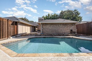 A rectangular swimming pool with a stone border, surrounded by a wooden fence and a brick building with a sloped roof.