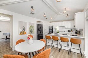 A modern kitchen with white cabinets, a black refrigerator, and a dining area with a white table and orange chairs.
