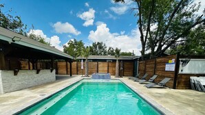 Una piscina rectangular con borde de azulejos, rodeada por una terraza de madera y una zona cubierta con techo.