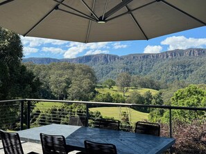 Un área de comedor al aire libre con una mesa, sillas, un paraguas grande y una vista panorámica de un valle con árboles y acantilados.