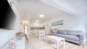 A modern living room with a grey sofa, a white coffee table, and a TV mounted on the wall. There is a kitchen area in the background with white cabinets and a dining table with chairs. - Casa Cris - Beach House By LVH (Puerto del Carmen)