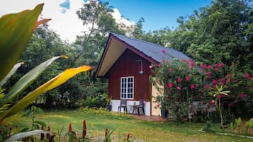 A small wooden house with a sloped roof, surrounded by lush greenery and flowering plants.