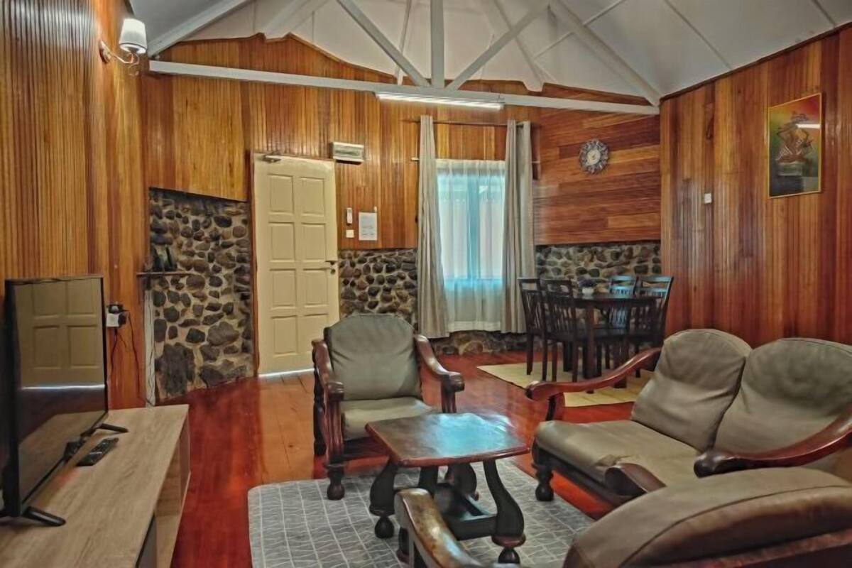 A cozy living room with wooden walls, a stone fireplace, a dining area with a table and chairs, and a large window with curtains.