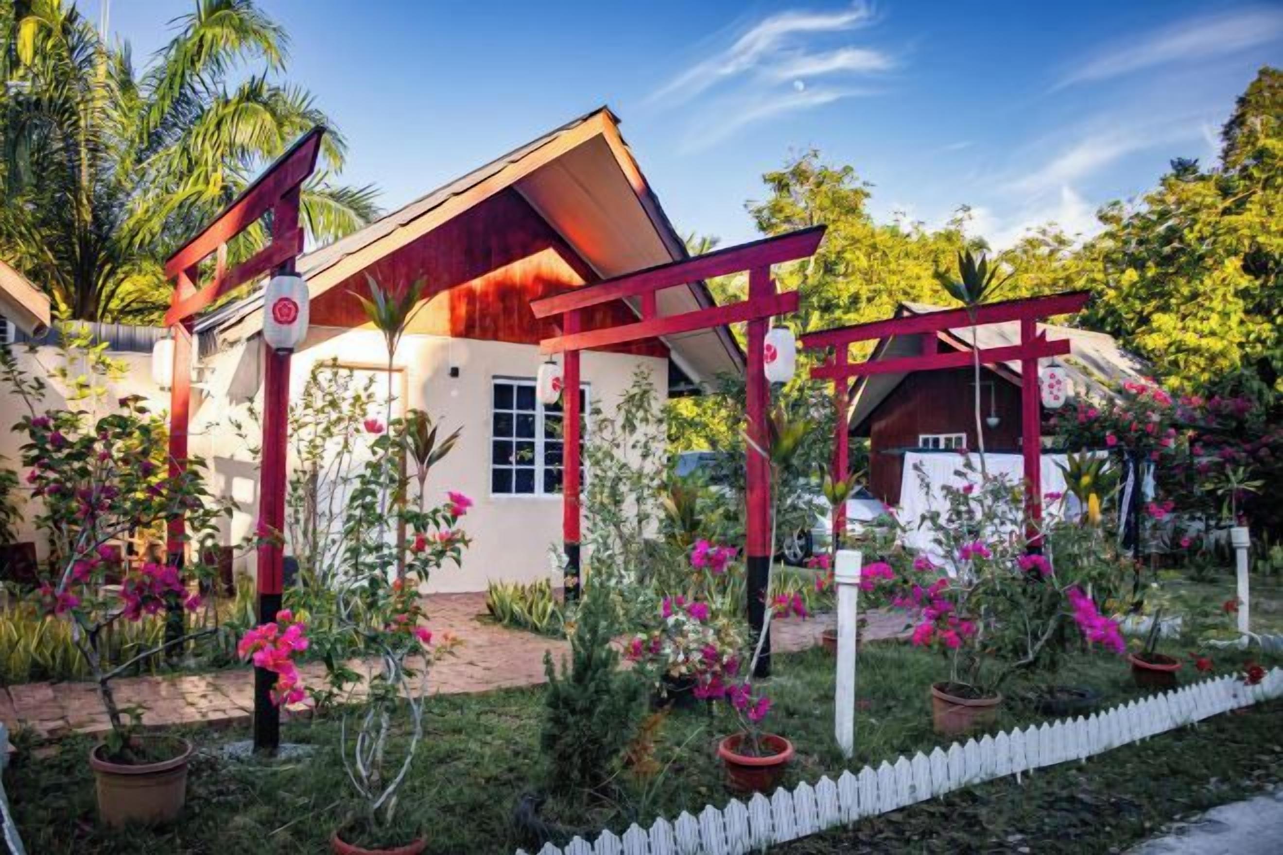A house with a red roof and white walls, surrounded by a garden with pink flowers and a white picket fence.