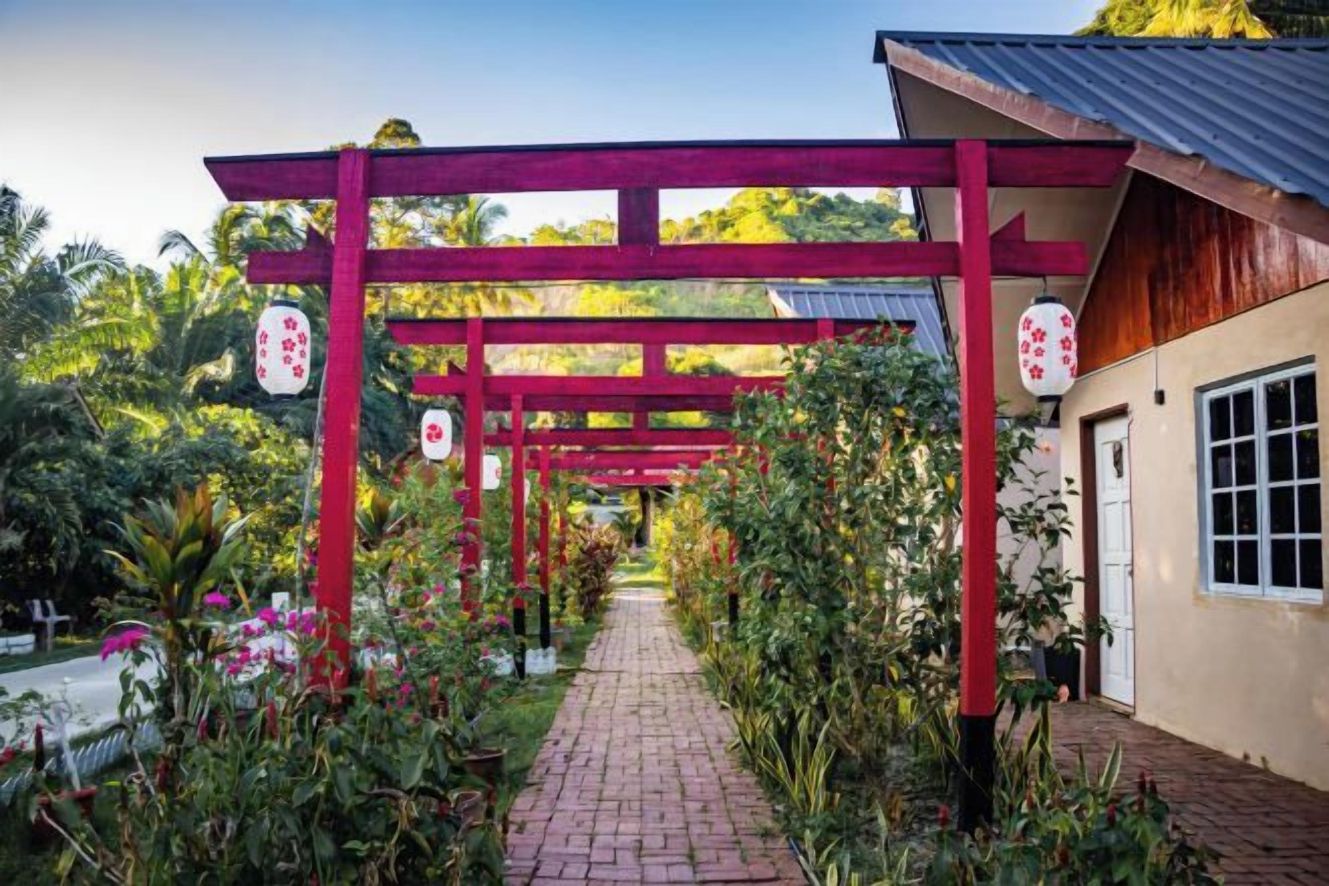 A pathway leading to a building with a red structure overhead, surrounded by greenery and plants.