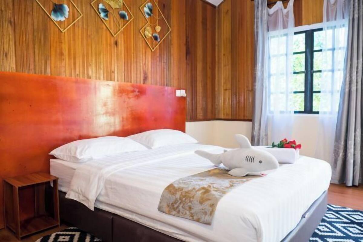 A hotel room with a wooden headboard, a bed with white linens, a plush white animal-shaped pillow, and a window with curtains.