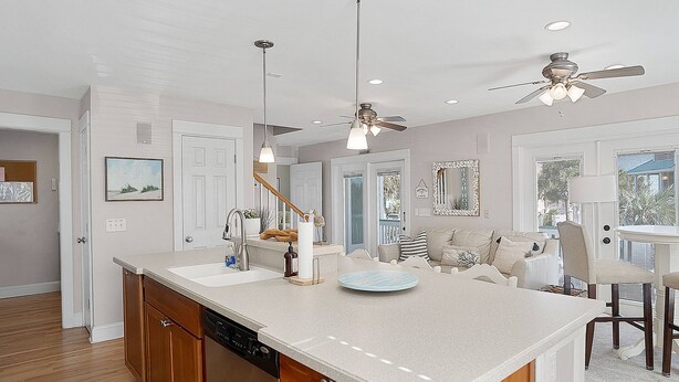 A spacious kitchen with wooden cabinets, a dishwasher, and a large island with a sink. There are ceiling fans, pendant lights, and a view of the living area through an open door. - Charming Ocean View Retreat- Steps to the Beach and Pool- Resort-Style Amenities (Saint Helena Island)