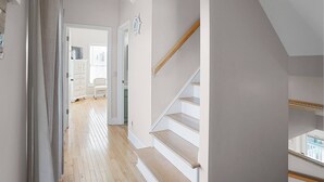 A hallway with wooden flooring, a staircase with white railings, and a door leading to a room with a white chair and a cabinet. - Charming Ocean View Retreat- Steps to the Beach and Pool- Resort-Style Amenities (Saint Helena Island)