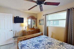 Un dormitorio con cama, cómoda de madera, televisor, ventilador de techo, ventana con cortinas y una silla.